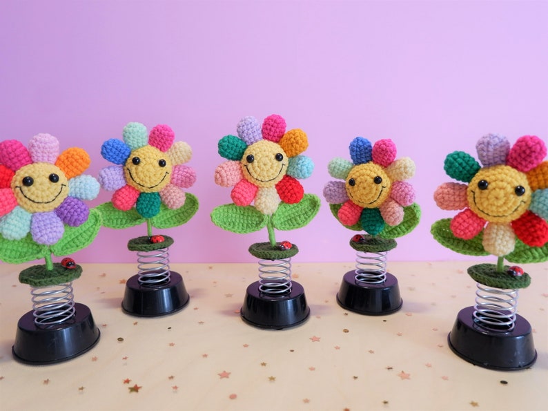 May include: Five colorful crocheted flowers with smiling faces on spring-loaded bases. Each flower has a different color combination of pink, purple, green, yellow, and blue.