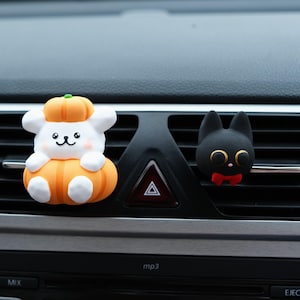Op de afbeelding: Twee auto-ventilatieopeningdecoraties. Eén is een wit stripfiguur met een pompoenhoed en zittend op een pompoen. De andere is een zwarte kat met een rode vlinderdas. Beide zijn bevestigd aan de ventilatieopening van de auto.
