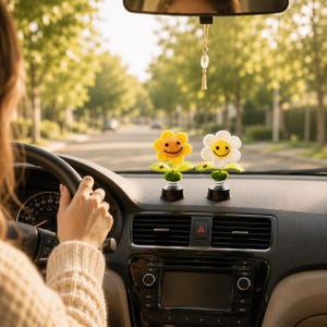 May include: Two smiling flower car decorations. One is yellow with an orange centre, and the other is white with a yellow centre. Both have green leaves and sit on black bases. The car's dashboard is visible, with a steering wheel and a road in the background.