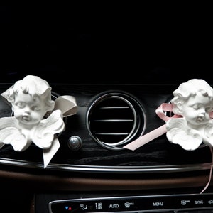 May include: Two white porcelain angel figurines with pink ribbon accents hanging from the air vents of a car dashboard.