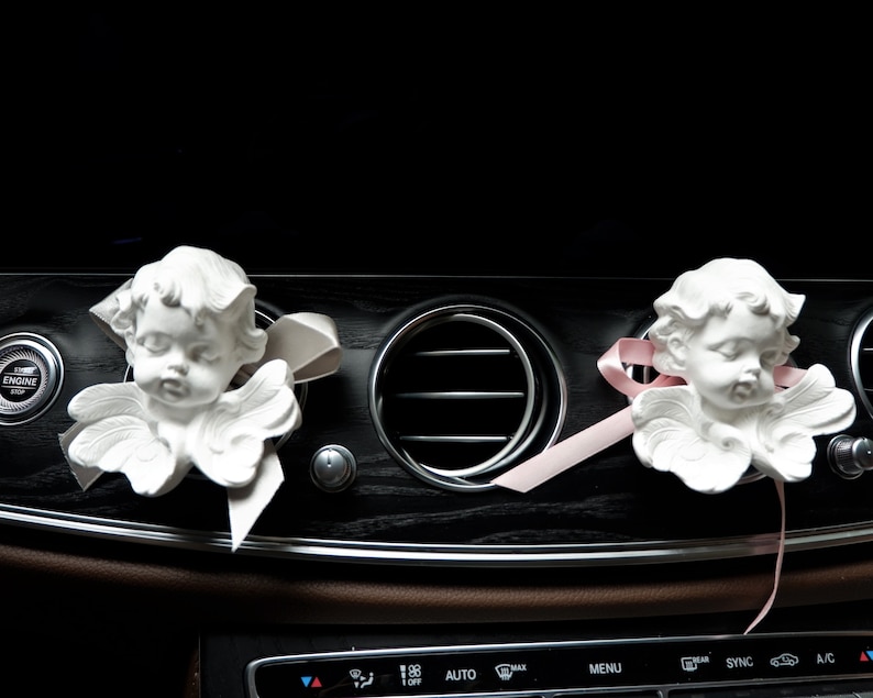 May include: Two white porcelain angel figurines with pink ribbon accents hanging from the air vents of a car dashboard.