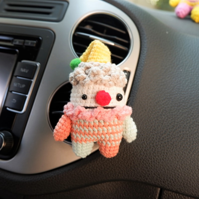 May include: A crocheted clown doll with a yellow hat, pink and green clothing, and a red nose. The doll is hanging from a car's air vent.