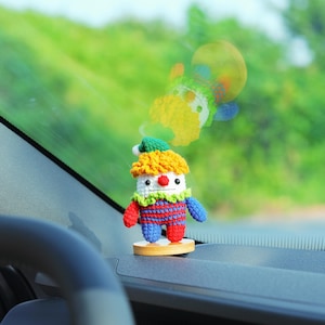 May include: A handmade crochet clown doll with a yellow wig, red nose, and a green and white collar. The clown is wearing a red and blue striped outfit and is mounted on a small wooden base. The background is blurred, showing a green, outdoor scene.