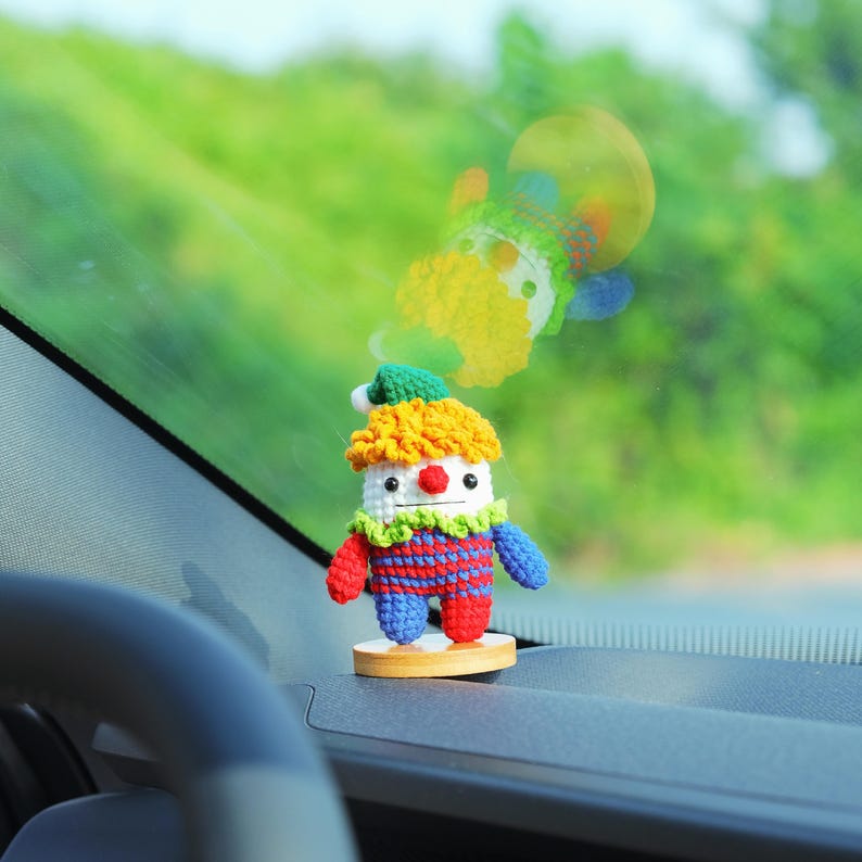 May include: A handmade crochet clown doll with a yellow wig, red nose, and a green and white collar. The clown is wearing a red and blue striped outfit and is mounted on a small wooden base. The background is blurred, showing a green, outdoor scene.