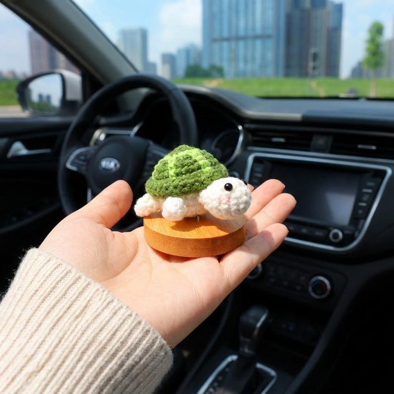 May include: A crocheted turtle figurine with a green shell and white body, mounted on a small wooden base. The turtle has black eyes and a pink mouth. The figurine is held in a hand, with a car interior in the background.