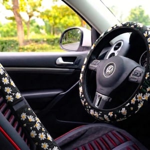May include: A black steering wheel cover and a matching seat belt shoulder pad, both featuring a repeating pattern of white daisy flowers with yellow centers. The accessories are installed on a black steering wheel and a black car seat belt.