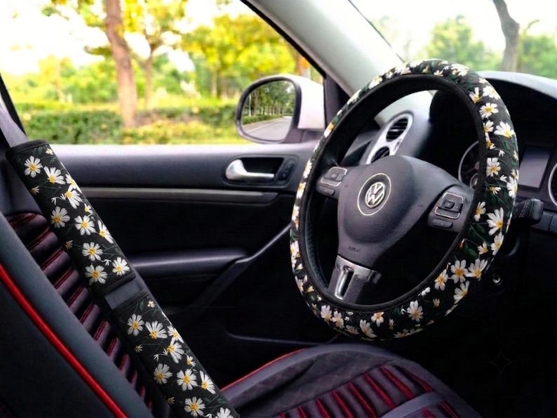 May include: A black steering wheel cover and a matching seat belt shoulder pad, both featuring a repeating pattern of white daisy flowers with yellow centers. The accessories are installed on a black steering wheel and a black car seat belt.