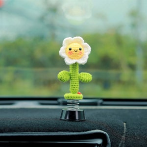 Könnte beinhalten: Eine gehäkelte Blume mit einem lächelnden Gesicht, gelber Mitte und weißen Blütenblättern, befestigt an einer Feder und einem schwarzen Sockel. Die Blume befindet sich auf einem schwarzen Armaturenbrett eines Autos.