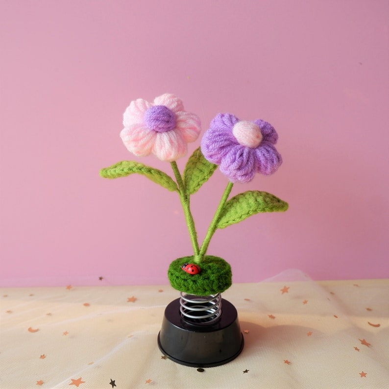 May include: A spring-loaded flower pot with two yarn flowers, one pink and one purple, with green leaves. A small red ladybug is on the green base.