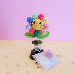 May include: A colorful crocheted flower with a smiling face sits on a spring-loaded base. The flower has green leaves and a red ladybug on the stem. The base is black with white polka dots.