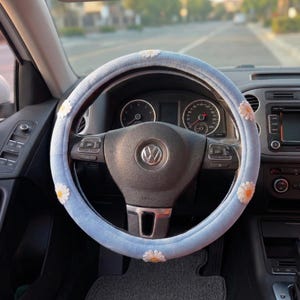 May include: A light blue steering wheel cover with white and yellow daisy accents. The cover is made of a soft, plush material and is designed to fit a standard car steering wheel. The car's dashboard and other interior components are visible in the background.