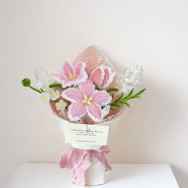 May include: A handcrafted bouquet of pink and white crocheted flowers, including lilies and smaller blossoms, wrapped in white paper and tied with a pink ribbon. A small card reads "I bought flowers thinking about you..."