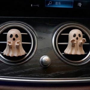 May include: Two light-colored wooden ghost figures with smooth, natural wood grain textures, black painted eyes, and open mouths. Each ghost has small arms extended forward and is attached to the circular air vent slats of a vehicle dashboard.