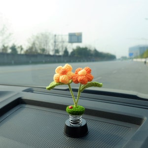 May include: A crocheted flower car air freshener with orange and yellow petals on a spring base. The flowers are attached to a green stem and are sitting on a black car dashboard.
