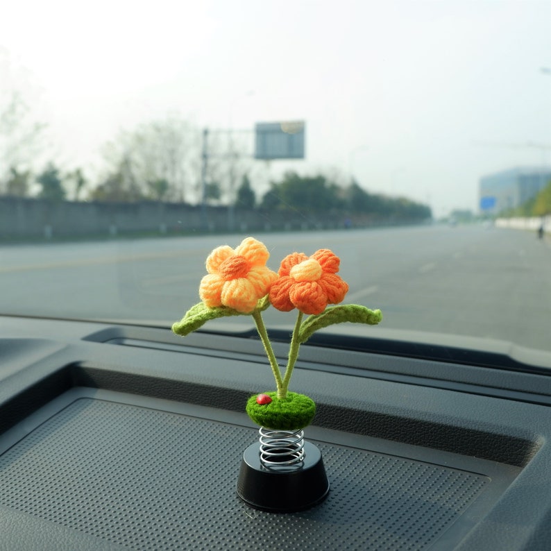 May include: A crocheted flower car air freshener with orange and yellow petals on a spring base. The flowers are attached to a green stem and are sitting on a black car dashboard.