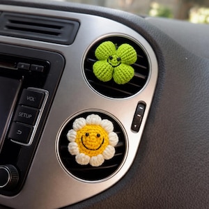 Op de afbeelding: Twee gehaakte auto-ventilatieclips met een felgroen klavertjevier en een wit met gele madeliefje. Beide items hebben geborduurde zwarte lachende gezichtjes met kleine zwarte kraaloogjes, bevestigd aan de lamellen van een dashboard.