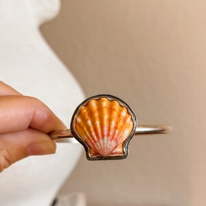 May include: A metal cuff bracelet featuring a central scallop shell ornament. The shell displays a gradient of orange, yellow, and white tones with distinct vertical ridges. The metal band is smooth and polished, providing a clean contrast to the textured, natural appearance of the shell centerpiece.