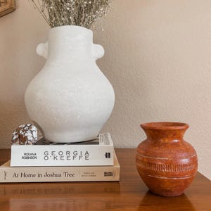 May include: A white ceramic vase with two small handles containing dried baby's breath flowers, placed on two stacked books titled Georgia O'Keeffe and At Home in Joshua Tree, next to a small orange textured clay pot and a small stone figurine.