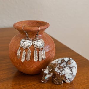 May include: A pair of silver earrings with a white center and feather dangles, displayed next to a small, brown and white bear figurine and a terracotta pot. The earrings are hanging from the pot.