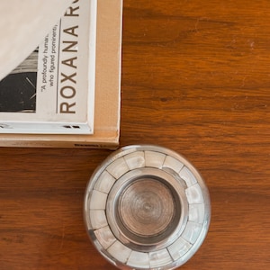 May include: A silver-toned, round candle holder with a decorative inlaid design of white rectangular pieces. The candle holder sits on a wooden surface. A book with the title "ROXANA R" is partially visible.