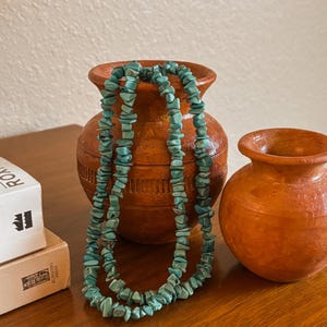 May include: A turquoise chip bead necklace draped over a brown terracotta vase. The necklace is made of irregular, light blue-green stones. A smaller vase and two books are also visible on the wooden surface.