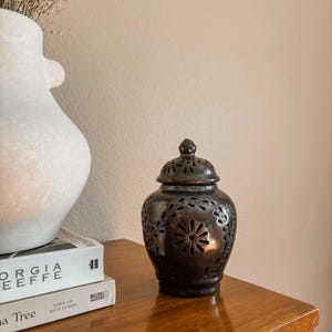 May include: A dark brown ceramic jar with a removable lid, featuring intricate cut-out floral patterns throughout the body and lid. The jar has a rounded silhouette with a small knob on top, allowing light to pass through the decorative openings.