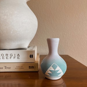 May include: A small, bulbous ceramic vase featuring a gradient color transition from light purple at the neck to a matte teal at the base. The lower portion is decorated with white, geometric, triangular patterns carved into the surface.