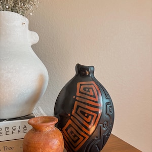 May include: A collection of decorative vases in various sizes and colors. A small, round, terracotta-colored vase sits next to a larger, black vase with an orange geometric design. A white vase with a unique shape is in the background.