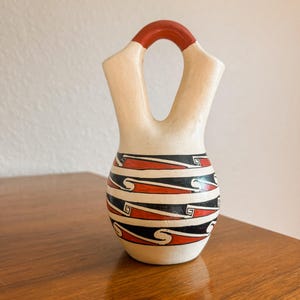 May include: A ceramic vessel with a cream-colored base, featuring a distinctive bridge handle at the top. The rounded body is decorated with horizontal bands of black and red geometric patterns, including repeating wave-like and spiral motifs.