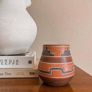 May include: A small, rounded terracotta vase featuring a matte finish with horizontal bands of dark gray and white geometric patterns. The vessel has a wide opening and a tapered base, showcasing a distinct contrast between the earthy orange clay and the dark, angular decorative motifs etched into the surface.