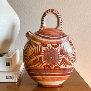 May include: A round ceramic vessel with a top handle and a small spout, featuring a cream-colored base with hand-painted reddish-brown floral and leaf motifs, horizontal stripes, and geometric patterns around the handle and body.
