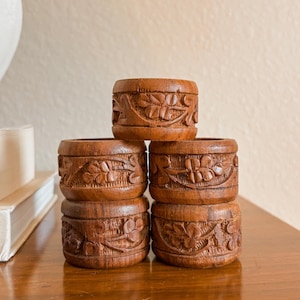 May include: A stack of six intricately carved wooden napkin rings. Each ring is a rich brown color and features a detailed floral and leaf design. The rings are stacked on a wooden surface, showcasing the craftsmanship.