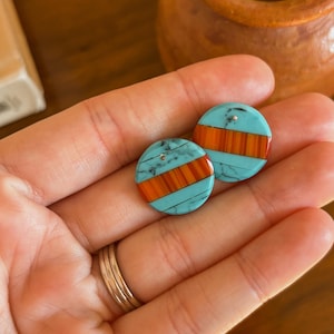 May include: Two round turquoise earrings with orange and brown stripes. The earrings have a small silver stud in the center. The earrings are held in a hand, with a blurred background of a wooden surface and a clay pot.