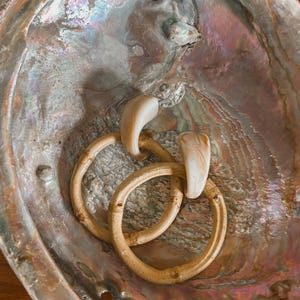May include: Abalone shell with iridescent colors, holding two bamboo hoop earrings and a bone-colored pendant. The earrings are light brown and have a natural, organic look. The shell's interior has a pearlescent sheen.