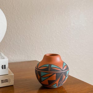 May include: A small, round, terracotta-colored clay vase featuring a matte finish. The exterior is decorated with a geometric pattern in black and teal, consisting of sharp, angular shapes and lines that wrap around the body of the vessel.