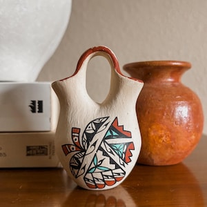 May include: A small, off-white ceramic vase with a handle, featuring a Native American-inspired design in black, red, and turquoise. The rim of the vase is painted red. A second, smaller, orange vase is in the background.