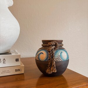 May include: A small, round, dark brown wooden vase featuring a decorative band with painted light blue and peach wave patterns and a cross-hatched design. A thick, natural-colored jute rope is tied around the neck of the vase with a knot.