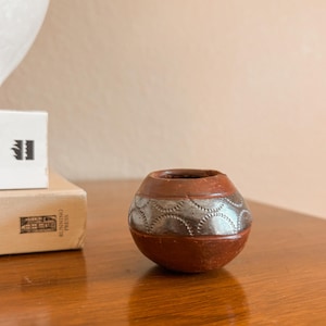 May include: A small, round, terracotta-colored clay pot featuring a wide, metallic silver band around its center. The silver band is decorated with a repeating pattern of stamped, semi-circular indentations along its top and bottom edges.