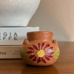 May include: A small, round, terracotta-colored clay pot featuring two small handles on the sides. The exterior is decorated with a hand-painted flower design, showcasing dark red petals surrounding a bright yellow center, accented by green leaves.