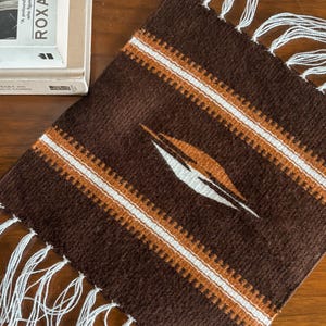 May include: A rectangular woven textile featuring a deep brown base with horizontal stripes in white and burnt orange. The center displays a stylized geometric diamond motif in white and orange. The top and bottom edges have white fringe.