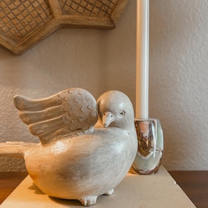 May include: A cream-colored ceramic dove figurine with detailed wing carvings, perched on a book titled "The Art & Life of Georgia O'Keeffe." A tall white candle and a decorative candle holder are also present.