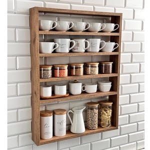 Op de afbeelding: Een rechthoekige houten wandplank met vijf horizontale niveaus. De planken bevatten diverse witte keramische mokken, doorzichtige glazen potten met houten deksels gevuld met granen en pasta, witte voorraadpotten met tekst en een witte keramische kan.