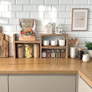 Può includere: Uno scaffale da cucina in legno a due livelli contenente barattoli di vetro con coperchi in legno, contenitori in ceramica bianca e due ornamenti bianchi a forma di casa. Lo scaffale è esposto su un piano di lavoro in legno con taglieri.