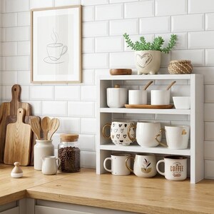 Op de afbeelding: Een wit houten rek met drie niveaus met diverse keramische mokken met hartjes- en bloemenpatronen, naast kleine witte potjes. De mokken hebben goudkleurige oren en op één mok staat de tekst Coffee Quality aan de zijkant.