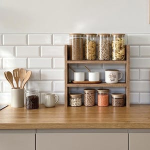 Op de afbeelding: Een houten aanrechtplank met drie niveaus met glazen potten met bamboedeksels gevuld met diverse droge waren, twee witte keramische mokken en een kleine kan. Houten keukengerei in een houder en twee houten snijplanken staan ernaast.