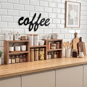 Op de afbeelding: Een aanrecht met twee houten planken met diverse glazen potten gevuld met droge waren, witte keramische bakjes en twee kleine huisvormige ornamenten. Het woord coffee is in zwarte schrijfletters op de wit betegelde muur bevestigd.