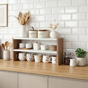 Op de afbeelding: Een houten en wit rek met twee niveaus met diverse keramische mokken, voorraadpotten en kleine schaaltjes. Links staan twee houten snijplanken en een houder met houten lepels. Een glazen pot met koffiebonen en een kleine plant in een pot staan ernaast.