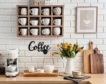 Può includere: Una rastrelliera da parete in legno per tazze con dodici scomparti contenenti varie tazze in ceramica, una macchina per espresso bianca, un vassoio in legno con teiera e tazzine, una brocca bianca con tulipani arancioni e due taglieri in legno.