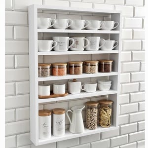 Puede incluir: Una estantería de cocina blanca montada en la pared, llena de tazas, tarros y recipientes blancos. La estantería muestra varios elementos esenciales de cocina, incluyendo tazas de café, tarros de especias y una jarra blanca. La unidad está montada en una pared de azulejos blancos.