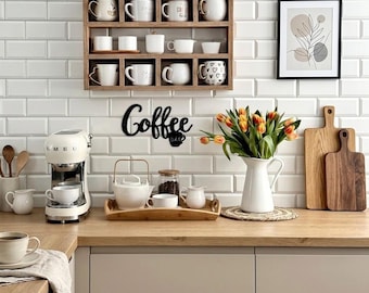 Può includere: Una mensola a muro in legno con varie tazze bianche, una macchina per espresso color crema, un'insegna in metallo nero con la scritta Coffee Bar, una brocca bianca con tulipani arancioni, un vassoio in legno con teiera e due taglieri.